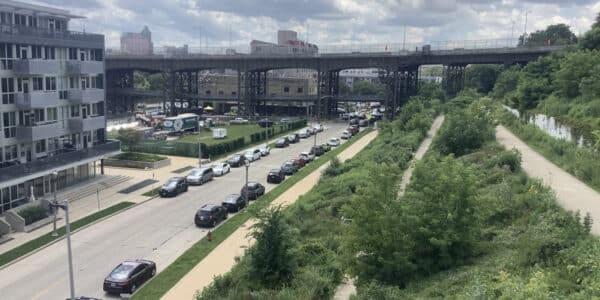 A line of cars parks along a city street beside a modern building, with a raised railway bridge and overgrown green space under a partly cloudy sky.