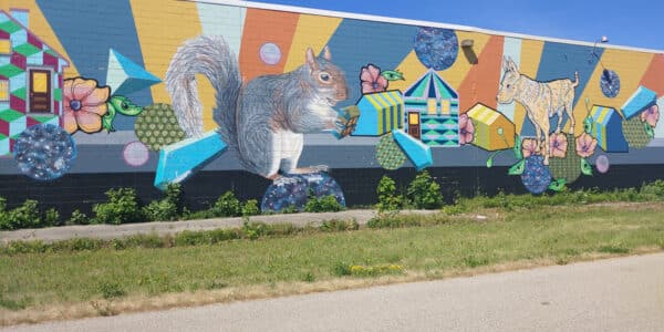 A colorful mural on a long brick wall features a large squirrel, a cat, geometric shapes, flowers, and abstract patterns under a clear blue sky.