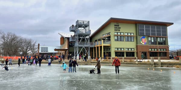 People ice skating outdoors on a frozen rink in front of a modern two-story building labeled Urban Ecology Center on a cloudy day.