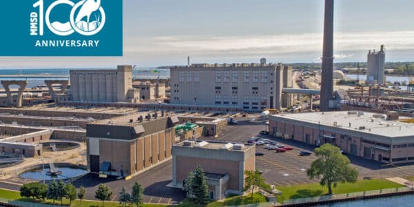 Aerial view of an industrial facility by the water with a tall smokestack; “100th Anniversary” logo appears in the upper left corner.