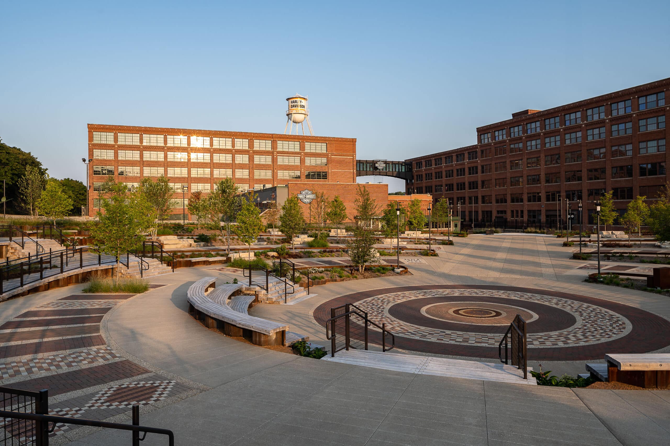 A large, open plaza with circular patterns, tiered seating, and young trees is surrounded by brick industrial buildings and a water tower in the background.