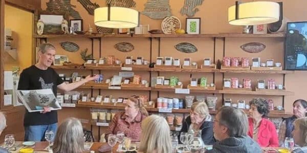 A man stands at the front of a room, speaking to a group of people seated at a table set for a tasting or class; shelves behind him display various packaged goods.
