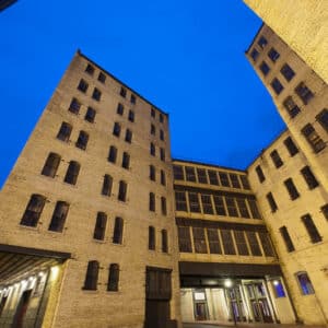 Tall brick buildings with many windows surround a paved courtyard under a deep blue evening sky, with a streetlamp visible in the foreground.
