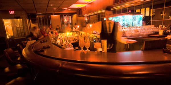 Dimly lit bar with a curved counter, leather stools, a bartender in motion, and cocktail glasses arranged on the counter and shelves.