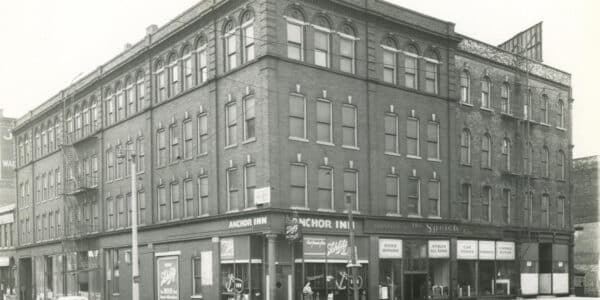 Black and white photo of a three-story brick building with businesses on the ground floor, including Anchor Inn and Spieth Co., cars parked along the street.
