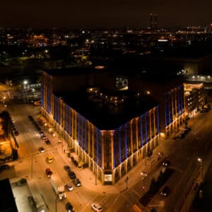 Aerial night view of a large, illuminated building with colorful vertical lights, surrounded by city streets and adjacent buildings.