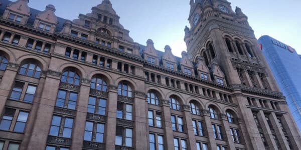 A historic multi-story building with arched windows and a tall clock tower, photographed from below against a clear blue sky. A modern glass building is visible in the background.