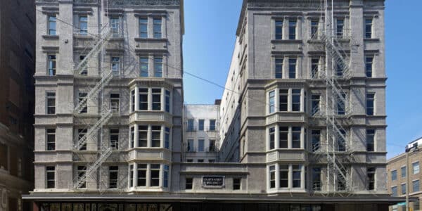 Historic six-story building with a central archway, fire escapes on both sides, retail stores at street level, and parked cars along the road in front.