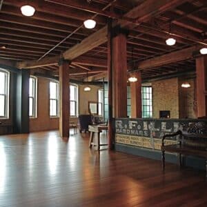 Spacious loft with exposed wooden beams, brick walls, large windows, wooden floors, vintage furniture, and a sign reading "R.F. LU Hardware.