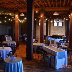 An event space with wooden beams, round and rectangular tables covered in white cloths, string lights, and wooden floors, set up for a gathering.