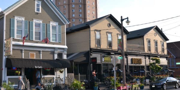 A street view shows several small businesses and restaurants in historic-style buildings, with outdoor seating, potted plants, and a tall modern apartment building in the background.