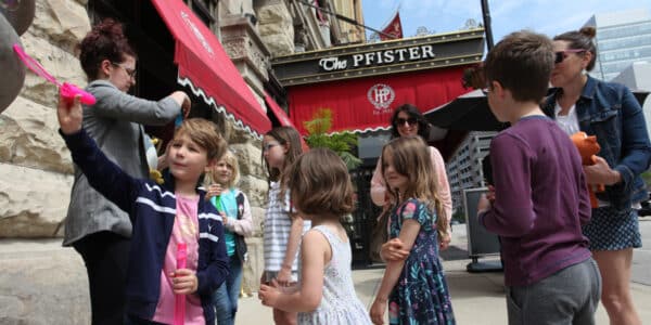 A group of children and adults stand outside The Pfister hotel, some holding bubble wands, on a sunny day.