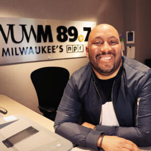 A man smiles while seated in a radio studio, with a sign reading "WUWM 89.7 Milwaukee's NPR" visible in the background.