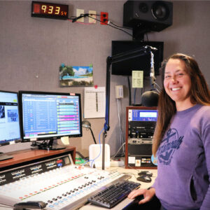 A woman stands smiling in a radio studio next to audio equipment, computer monitors, and a microphone. The wall clock reads 9:33.