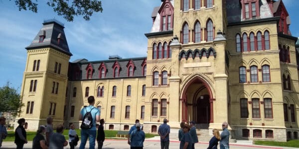 A group of people stands and sits outside a large historic building with arched windows and towers on a sunny day.