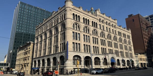 A large, historic stone building with arched windows stands at a city intersection on a clear day, surrounded by taller modern and older structures.