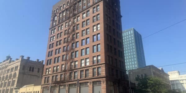 A tall, red-brick historic building with a fire escape on the facade stands on a city street under a clear blue sky.