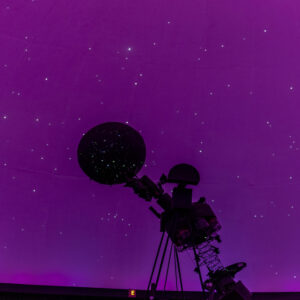 Silhouette of a planetarium projector against a purple-lit dome with simulated stars.