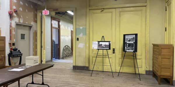 A room with yellow doors, wooden floors, a long table with documents, a black rotary phone, and two easels displaying black-and-white photographs. An open doorway leads to another hallway.