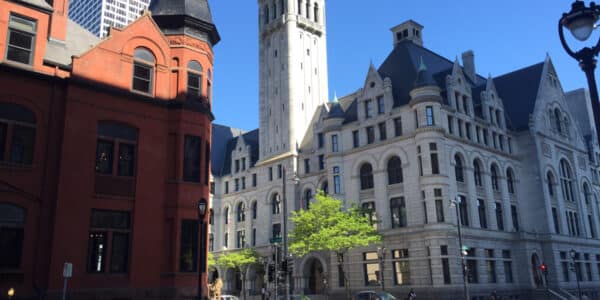 A red brick building stands beside a large gray stone historic building with a tall clock tower under a clear blue sky in an urban setting.