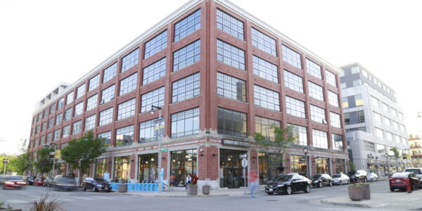 A large, modern brick building with many windows stands on a city street corner. Cars are parked along the street and pedestrians are walking nearby.
