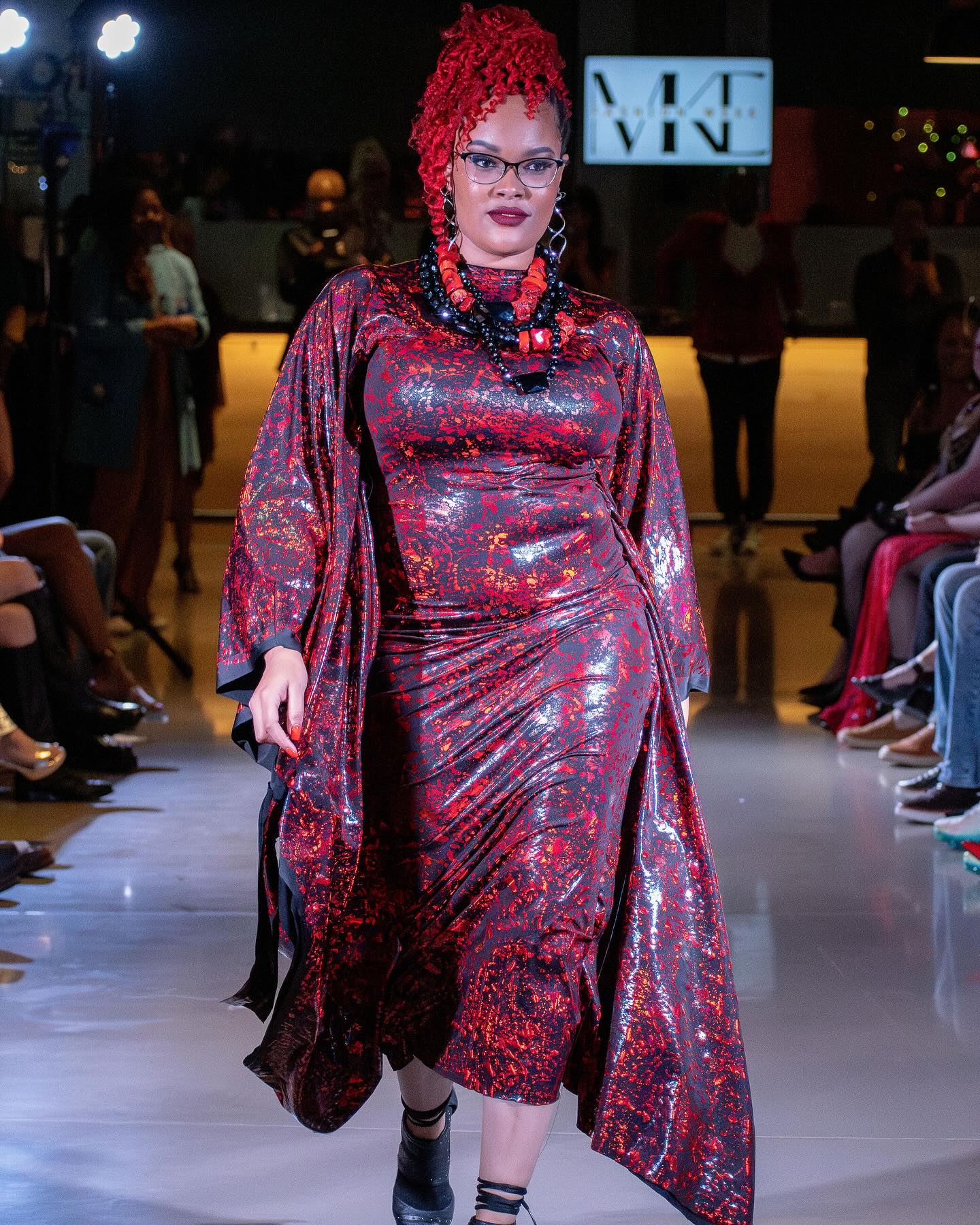 A model with red hair and glasses walks a runway wearing a shiny red and black patterned dress and statement necklace at a fashion show.