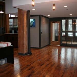 Modern office interior with wooden floors, exposed beams, hanging lights, a glass entry door, and cleaning supplies on a counter.