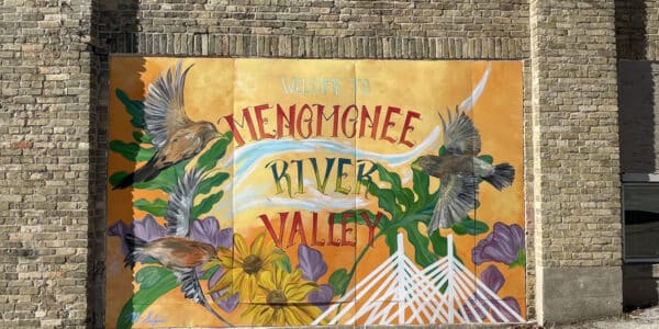 Mural on a brick wall reads "Welcome to Menomonee River Valley" with flying birds, purple flowers, green plants, and stylized white structures on a yellow background.
