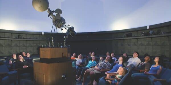 A group of people sit in a planetarium, looking up at a projection dome, with a large projector in the center of the room.