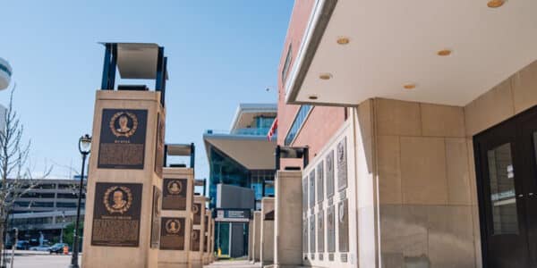 Outdoor walk with commemorative plaques mounted on pillars beside a building; modern stadium structure visible in background under a clear sky.