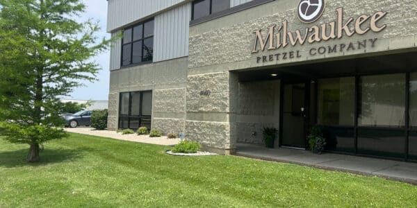 Exterior of the Milwaukee Pretzel Company building with a sign, large windows, and a grassy lawn with a small tree in front.