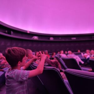 A group of people sit in reclining seats inside a planetarium with purple lighting; a boy in the foreground gestures with his hands.