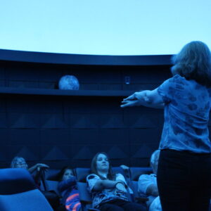 A woman stands at the front of a darkened planetarium, leading a group of seated people in a hands-on activity under a blue-lit dome.