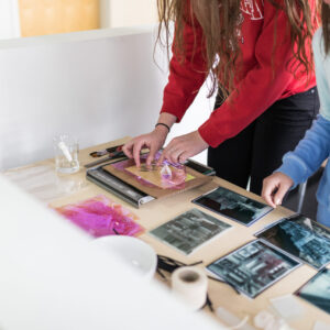 Two people working with photographs and art materials on a table, arranging prints and clear sheets for an art or craft project.