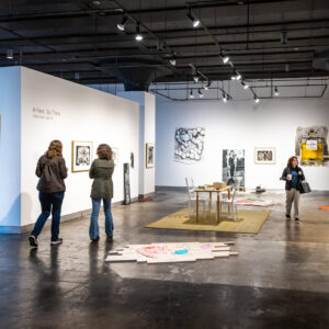 People view artwork displayed on white walls in a modern art gallery; various paintings, prints, and installations are visible, with three individuals observing the exhibits.