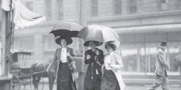 Three women wearing long skirts and hats walk in the rain with umbrellas on a city street; a man and a horse-drawn carriage are visible in the background.