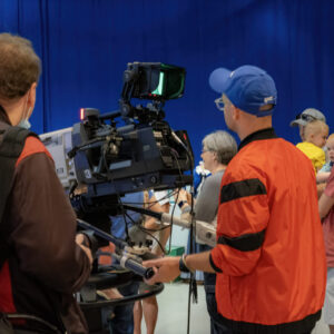 A camera crew films a group of people, including families with children, in front of a blue backdrop.