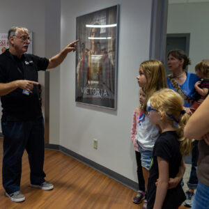 A man gestures while speaking to a group of women and children standing in a hallway next to a poster on the wall.