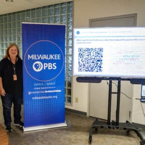 A person stands next to a Milwaukee PBS banner and a screen displaying a word cloud about Milwaukee PBS in an office setting.