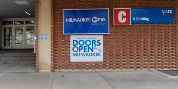 Entrance to a building with signs for Milwaukee PBS, C Building, MATC, and Doors Open Milwaukee, with a glass door and steps on the left.