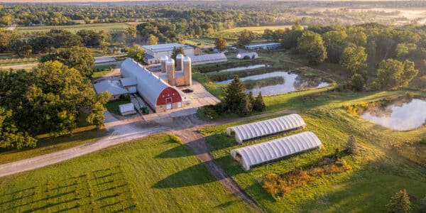 Aerial view of a farm with a red barn, silos, green fields, ponds, trees, and two white hoop houses at sunrise.