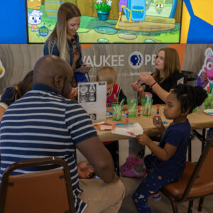 Adults and children sit at a table doing arts and crafts, with colorful cartoon fish decorations and a large screen in the background.