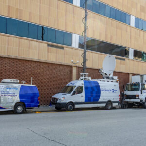 Three Milwaukee PBS broadcast vehicles, including two vans with satellite dishes and a large trailer, are parked on a city street beside a modern building.