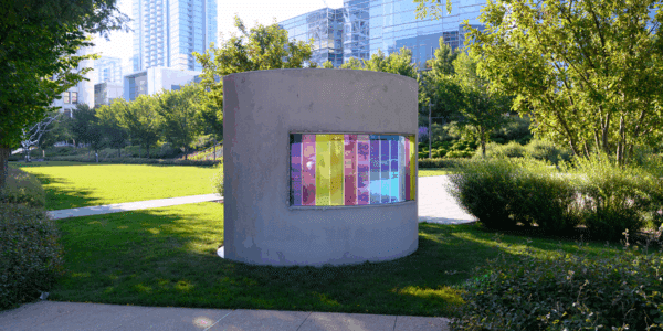A cylindrical concrete structure with a window displaying vertical strips of colorful translucent panels stands in a park surrounded by trees and modern buildings.