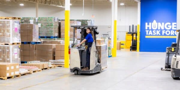 A person operates a forklift in a large warehouse with stacked pallets, near a blue wall labeled "Hunger Task Force.