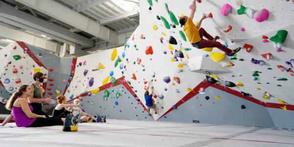 Three people watch as a climber ascends a colorful indoor bouldering wall in a brightly lit climbing gym.