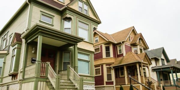 Three Victorian-style houses with porches and steep roofs stand side by side on a grassy slope under an overcast sky.