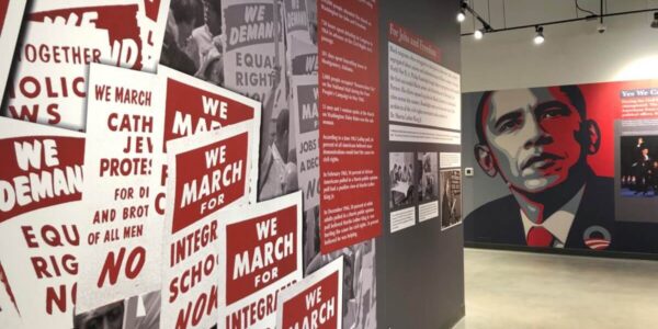 A museum exhibit features civil rights protest signs on one wall and an illustrated portrait of Barack Obama on another wall under bright lighting.