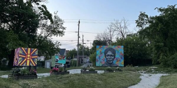 Three colorful outdoor murals, including a portrait of a woman, are displayed on a grassy roundabout surrounded by trees and small houses under a clear sky.