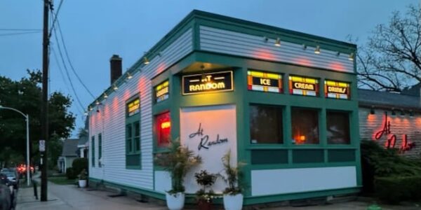 Small retro-style building with green trim at dusk, stained glass windows displaying “At Random” and “Ice Cream Drinks,” potted plants near entrance, street and houses visible in background.